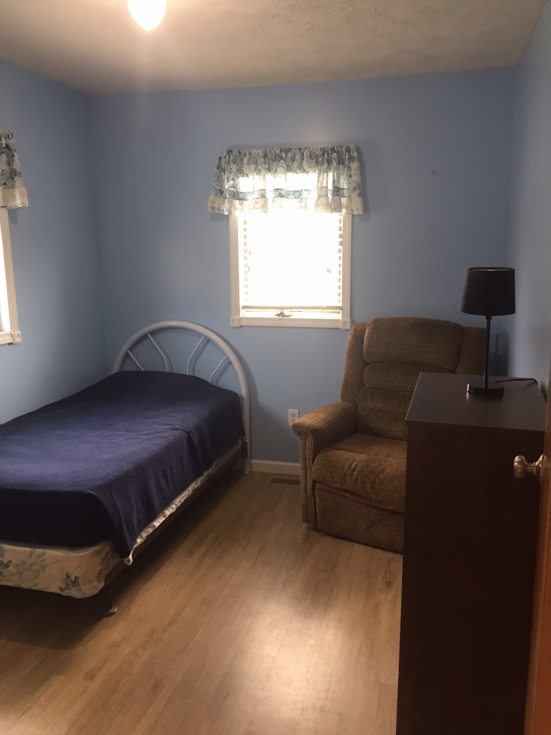 A small bedroom with light blue walls and wood flooring. The room contains a single bed with a dark blue blanket, a brown upholstered recliner chair, a wooden dresser with a black lamp on top, and two windows with floral valance curtains.