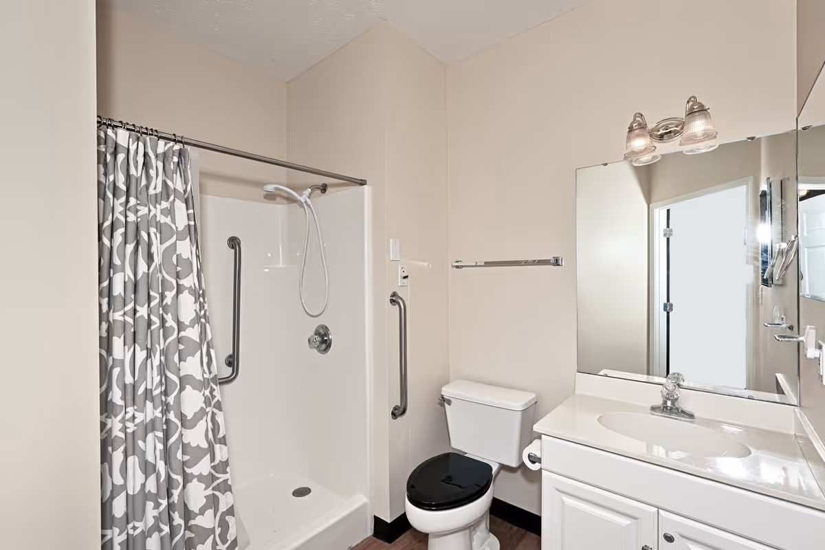 A clean bathroom featuring a white shower with a gray and white patterned shower curtain, a white toilet with a black seat, a white vanity with a sink, a large mirror above the sink, and a two-light fixture mounted on the wall above the mirror.