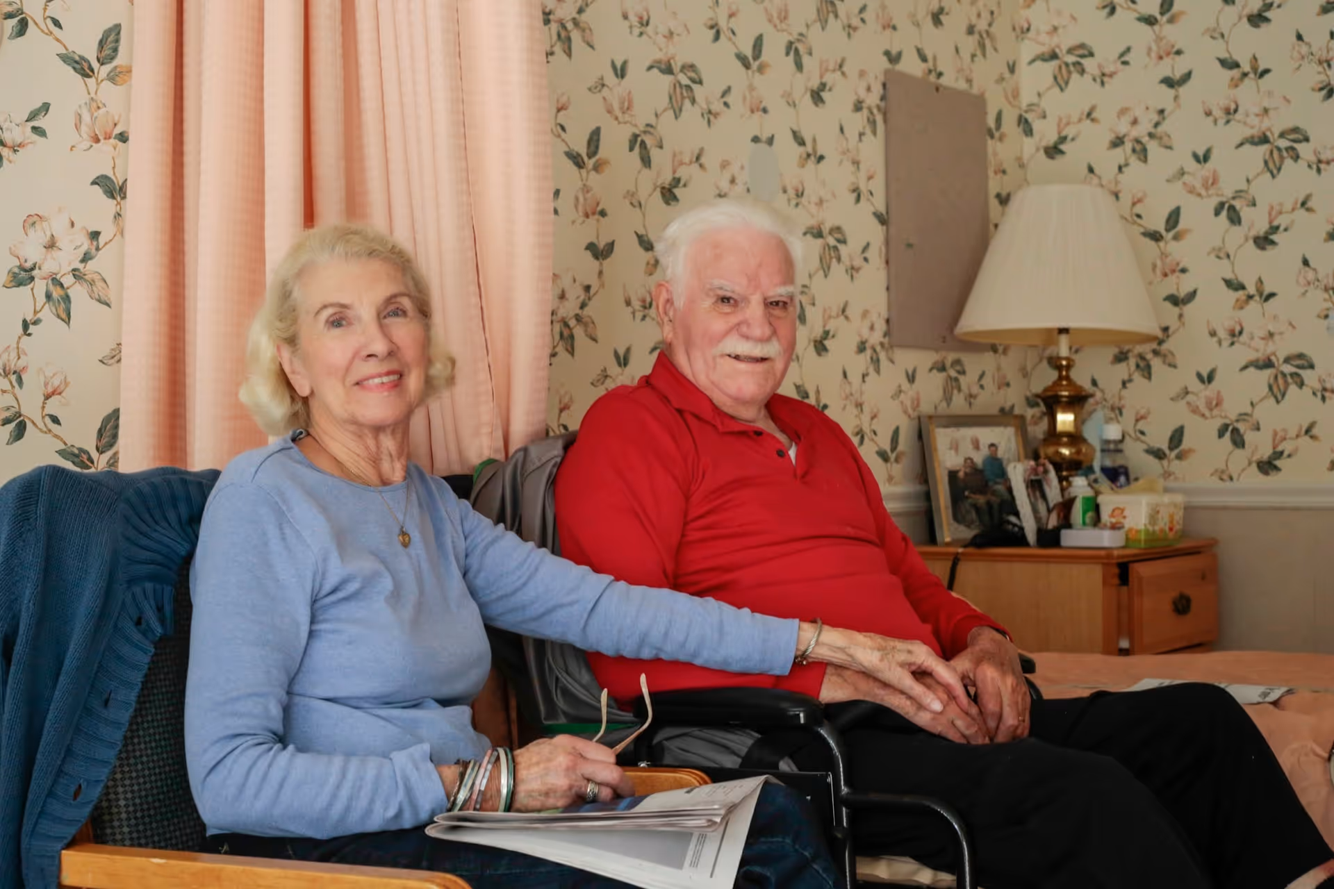 An elderly couple sitting together in a cozy room with floral wallpaper and a pink curtain. The woman is wearing a light blue sweater and holding a newspaper, while the man is in a red shirt sitting in a wheelchair. A wooden nightstand with a lamp, framed photos, and personal items is visible in the background.