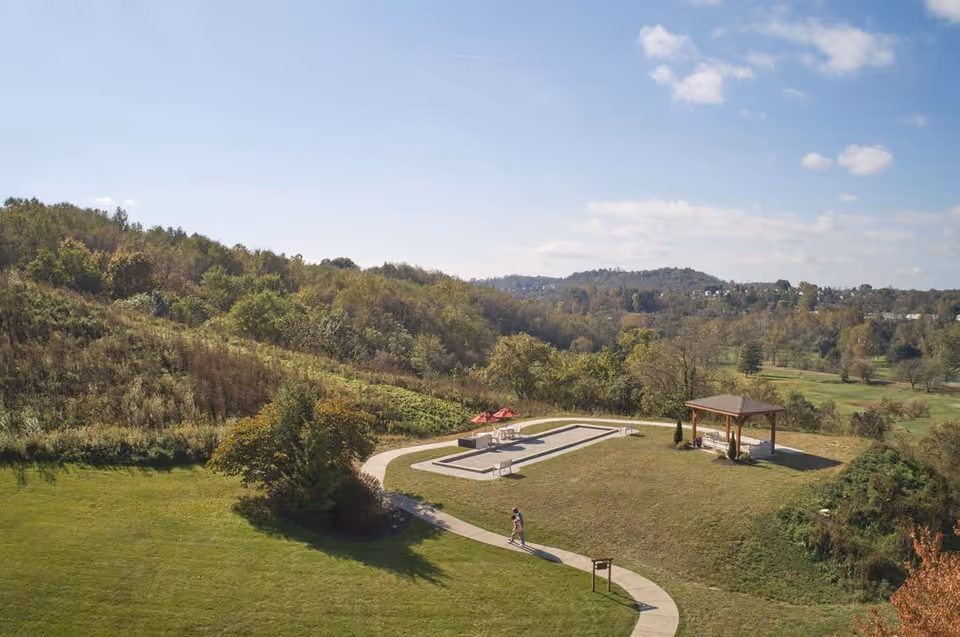 A scenic outdoor area at Friendship Village of South Hills featuring a walking path, a bocce ball court, a small gazebo, and surrounding greenery with hills and trees under a partly cloudy sky.