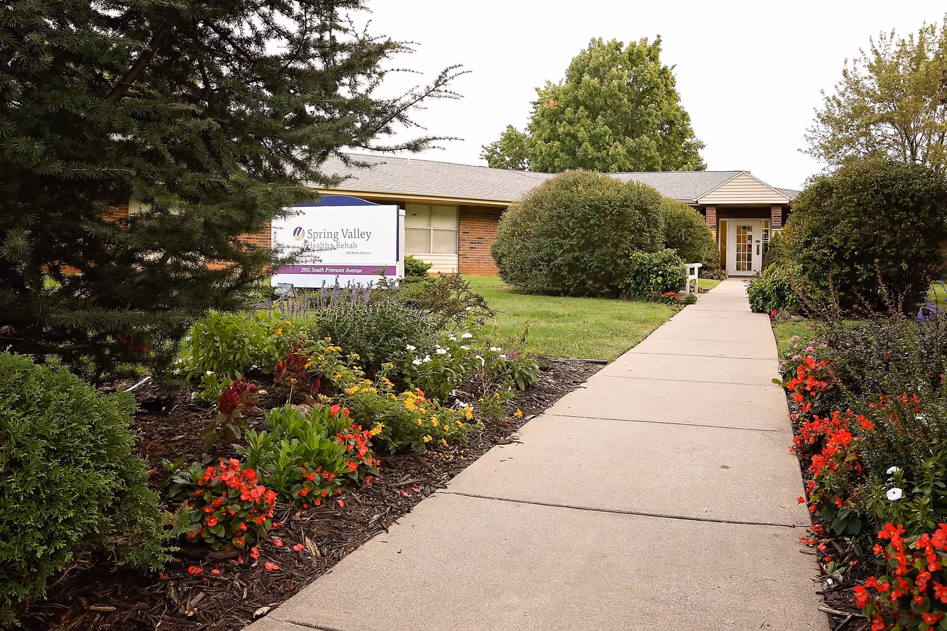 A concrete walkway leading to the entrance of a single-story brick building surrounded by well-maintained bushes and colorful flower beds. A sign near the walkway reads 'Spring Valley Health & Rehab' with the address 2915 South Fremont Avenue.
