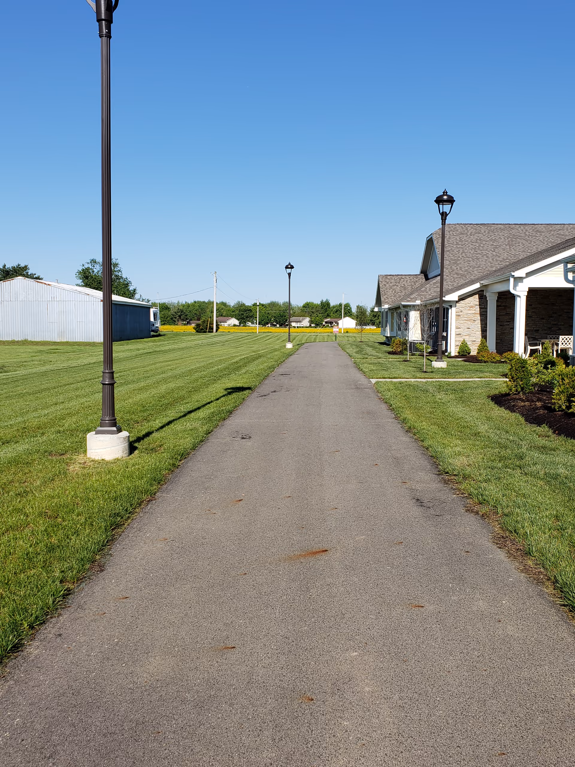A paved walkway flanked by green grass and lamp posts on both sides, leading towards a building with a sloped roof on the right and a large metal shed on the left under a clear blue sky.