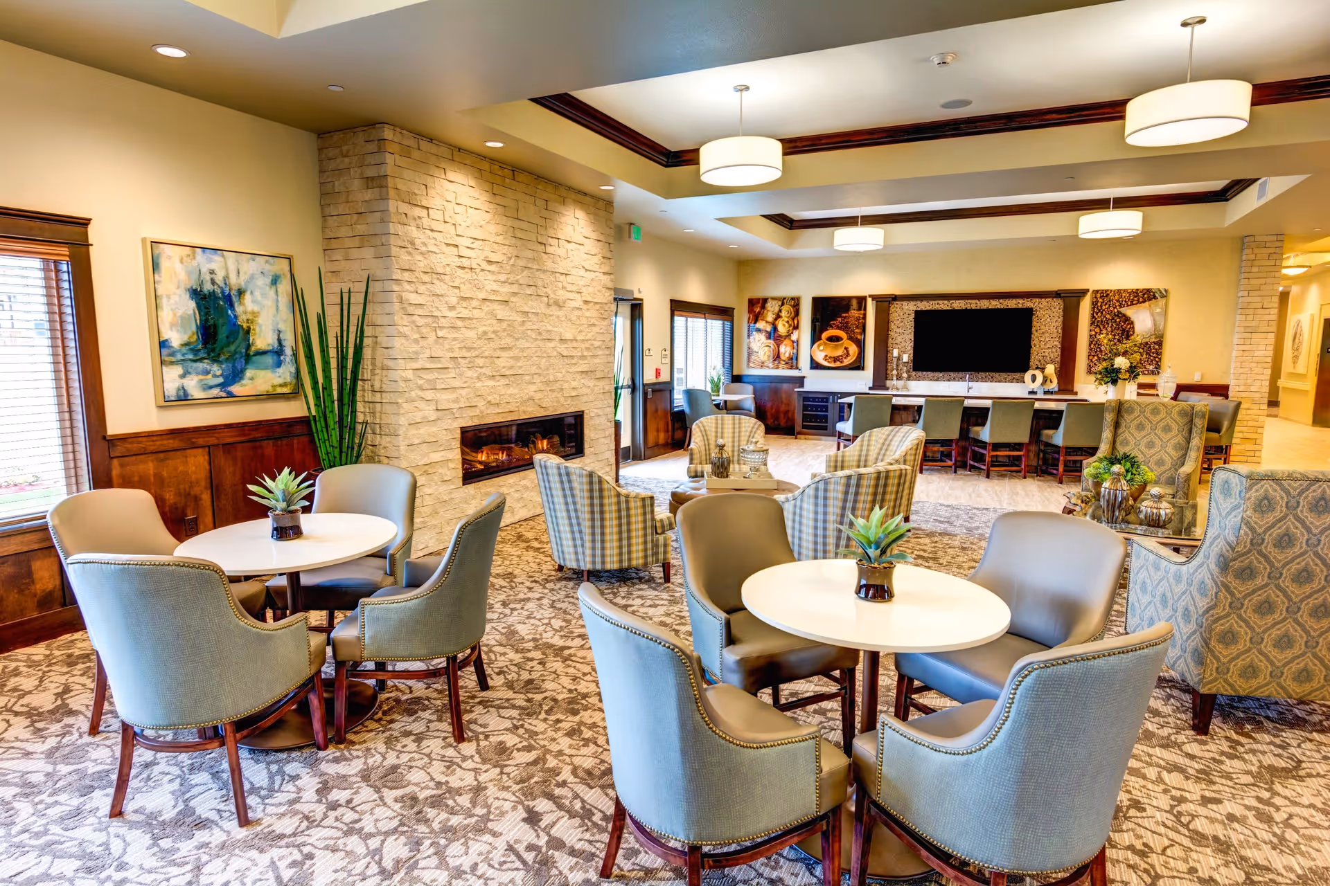 A spacious and well-lit senior living common area with multiple round tables surrounded by comfortable upholstered chairs. There is a stone fireplace on the left wall, abstract artwork, and large windows with wooden blinds. The back wall features a long counter with bar stools, decorative coffee-themed artwork, and a large flat-screen TV. The room has a patterned carpet and modern ceiling lights.