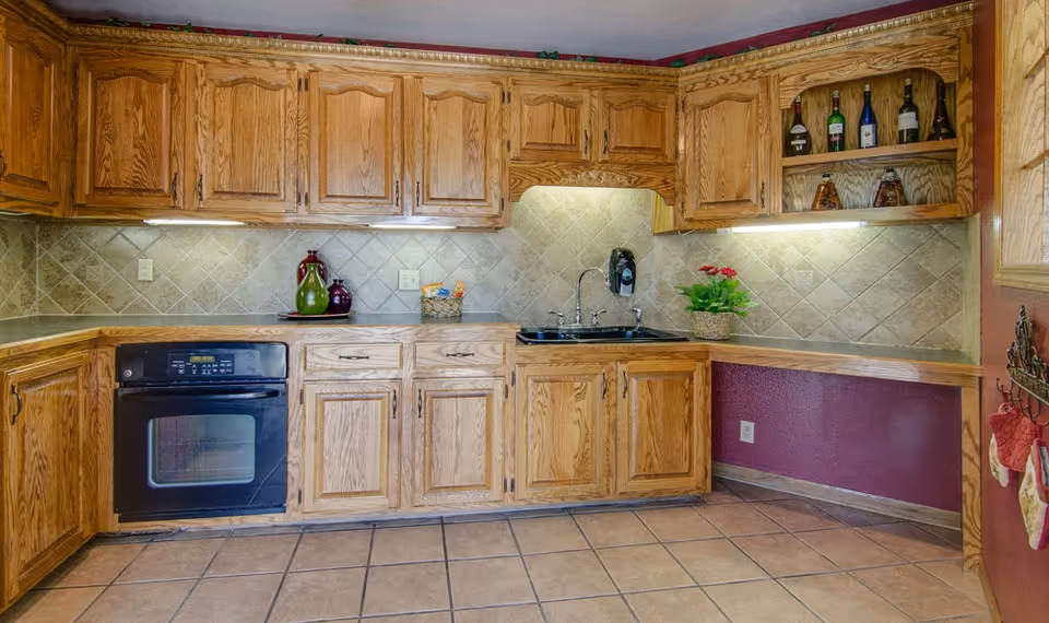 A kitchen with wooden cabinets and drawers, a black oven, a black sink with a faucet, and a tiled backsplash. There are decorative bottles and a small plant on the countertops. The walls are painted a reddish color and the floor is tiled.