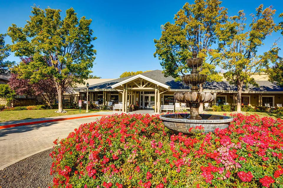 Front entrance of a senior living building with a multi-tier fountain surrounded by red flowers, trees, and a covered porte-cochère.