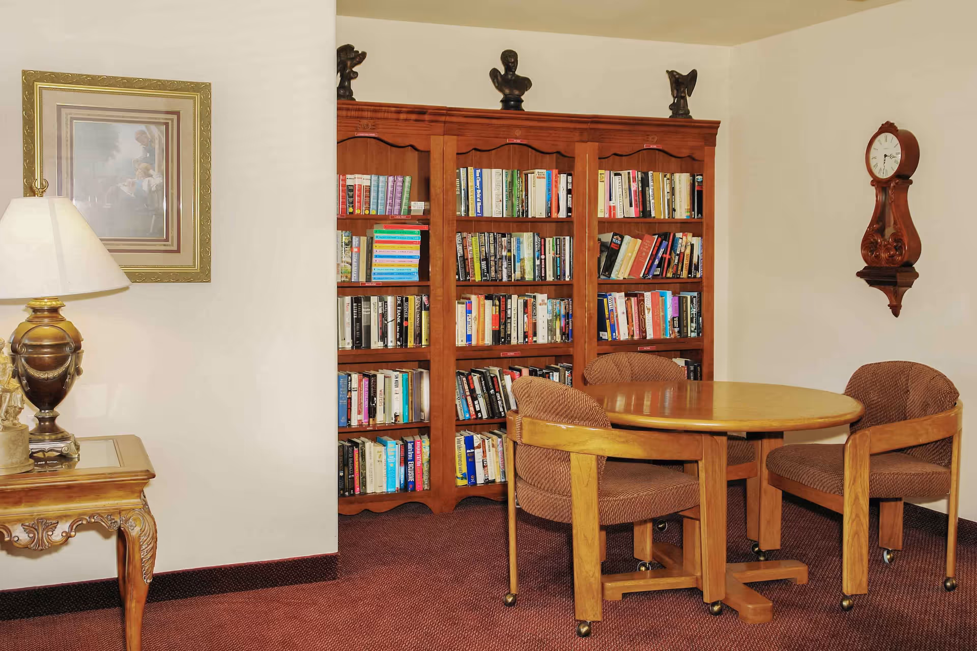 Small sitting area with a round wooden table and upholstered chairs in front of a large bookshelf, with a lamp, framed picture, and wall clock on the walls.