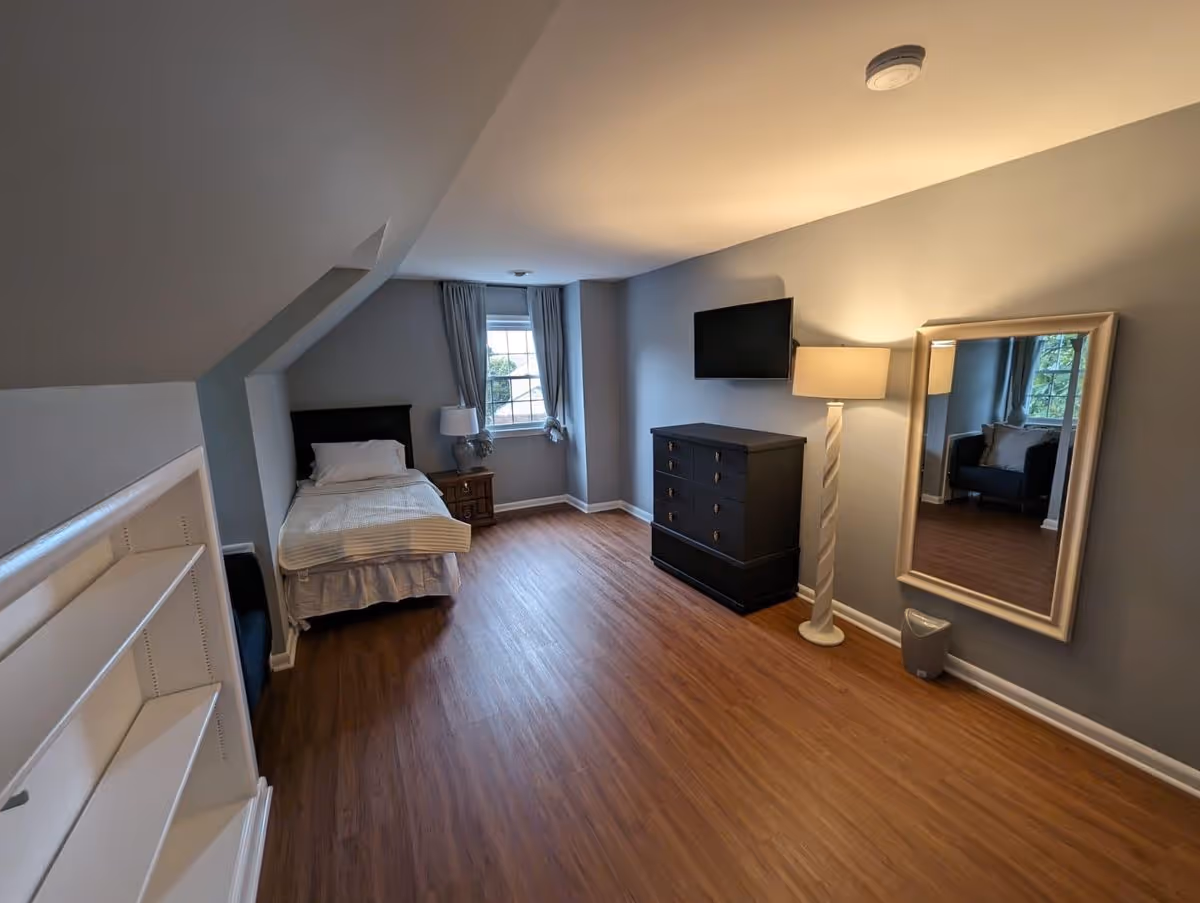 Spacious bedroom with a single bed by a window, dresser with wall-mounted TV, floor lamp, large mirror, and wood-look flooring.