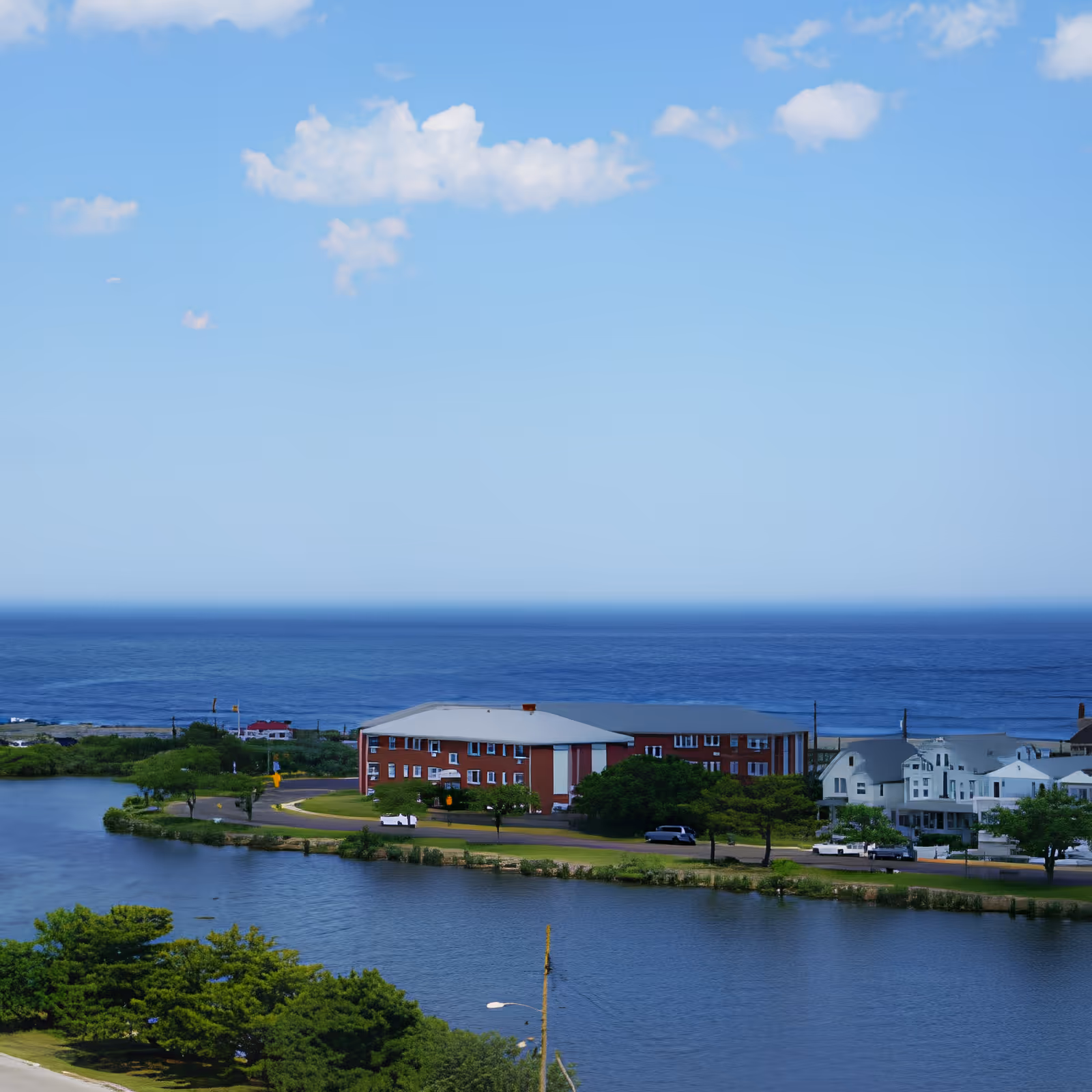 A scenic view of a waterfront area with a large body of water in the foreground, a red brick building and white buildings along the shoreline, and the ocean in the background under a blue sky with a few clouds.