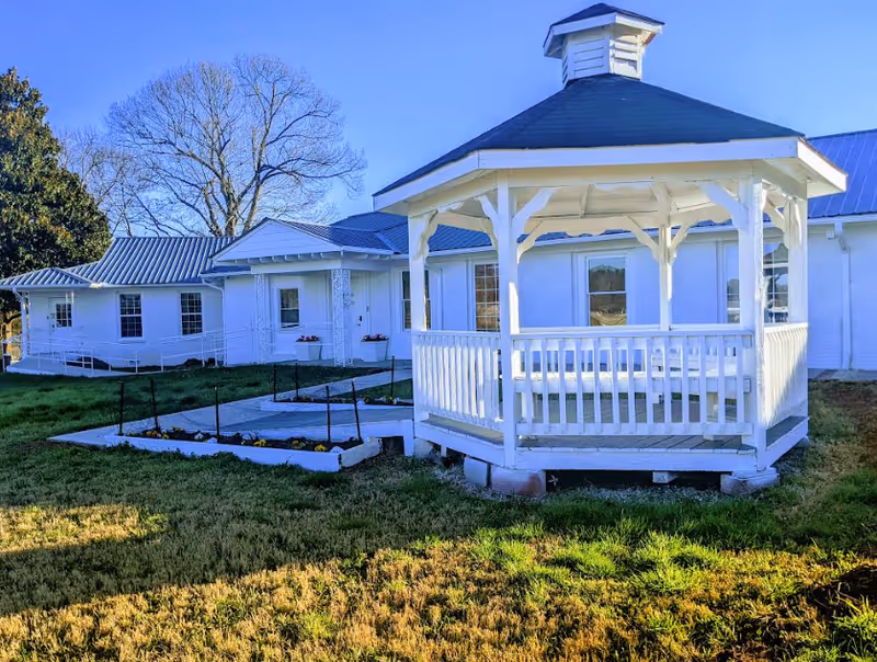 White wooden gazebo on a grassy lawn in front of a single-story white building under a clear blue sky.
