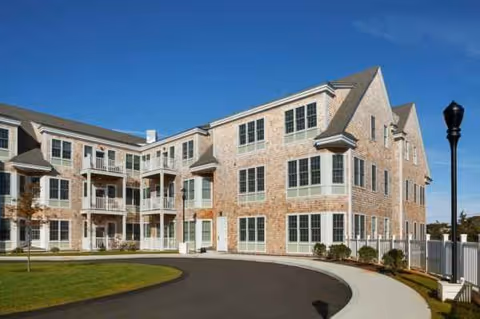 Brick senior living building with balconies, windows, and a curved driveway under blue sky.