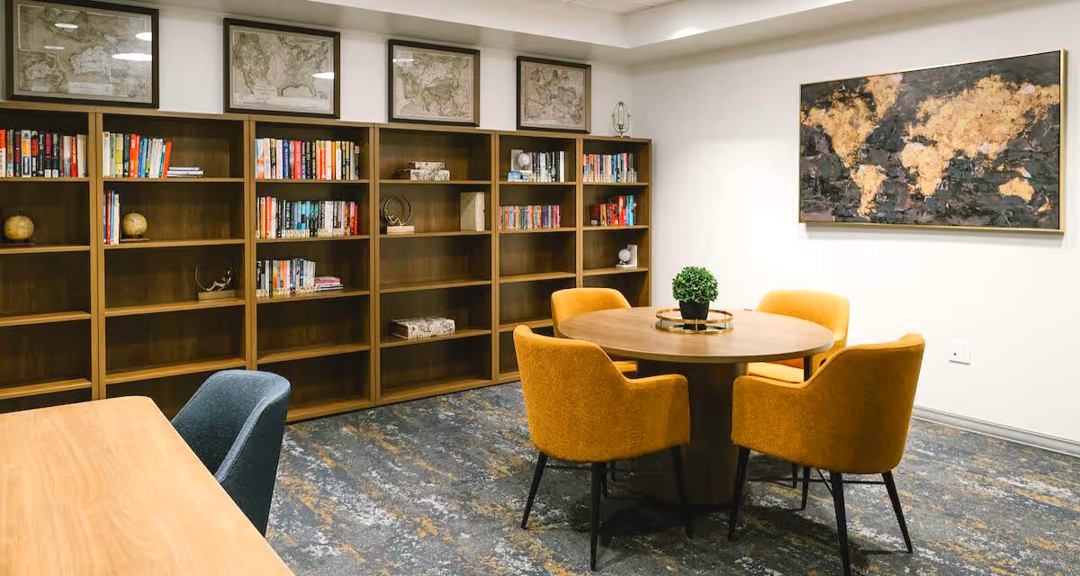 A communal reading room with wooden bookshelves, a round table surrounded by mustard-yellow chairs, and map artwork on the walls.