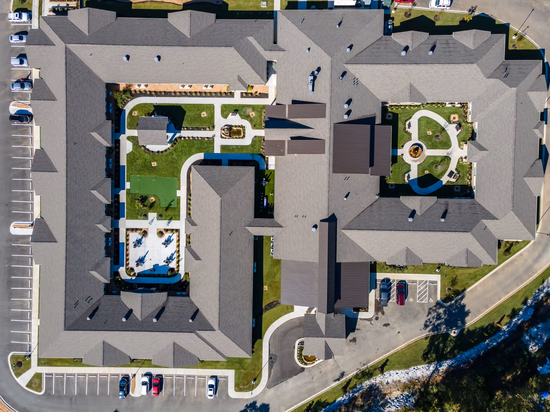 Aerial view of Crestview Senior Living facility showing the building's roof, landscaped courtyards with walking paths, seating areas, and parking lots surrounding the building.