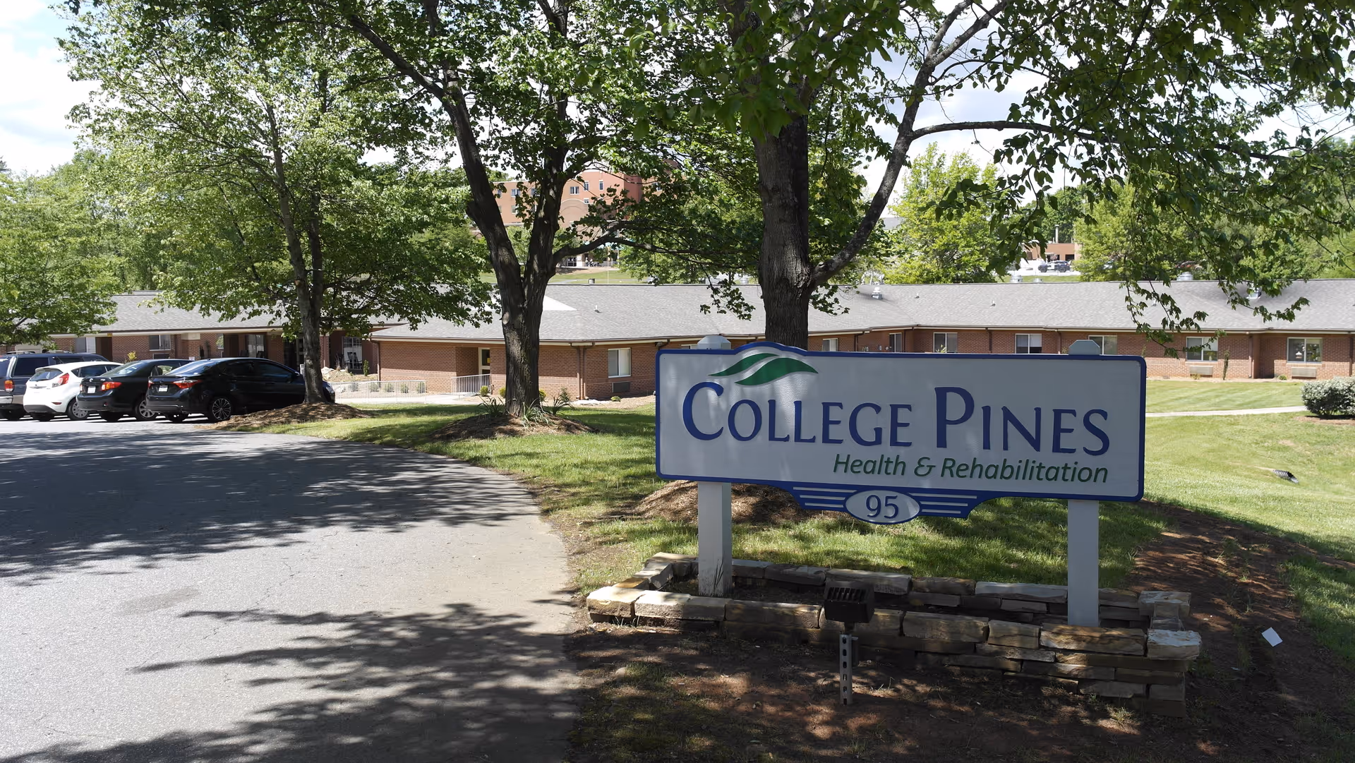 Entrance sign reading "College Pines Health & Rehabilitation" on a lawn in front of a single-story brick facility with trees and parked cars.