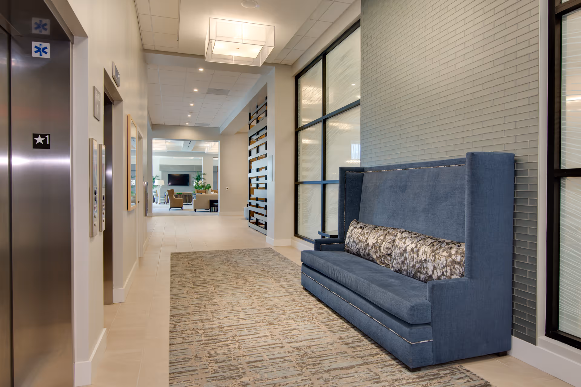 Modern interior lobby hallway with an elevator on the left and a blue high-backed sofa along a tiled wall.