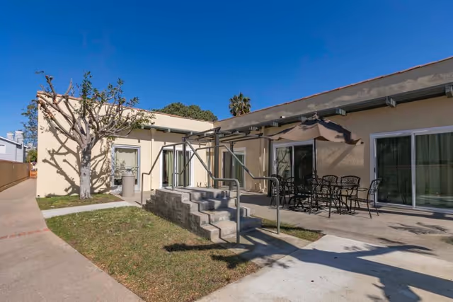 Outdoor courtyard and patio of a single-story facility with steps and a wheelchair ramp, patio table and umbrella, and sliding glass doors.