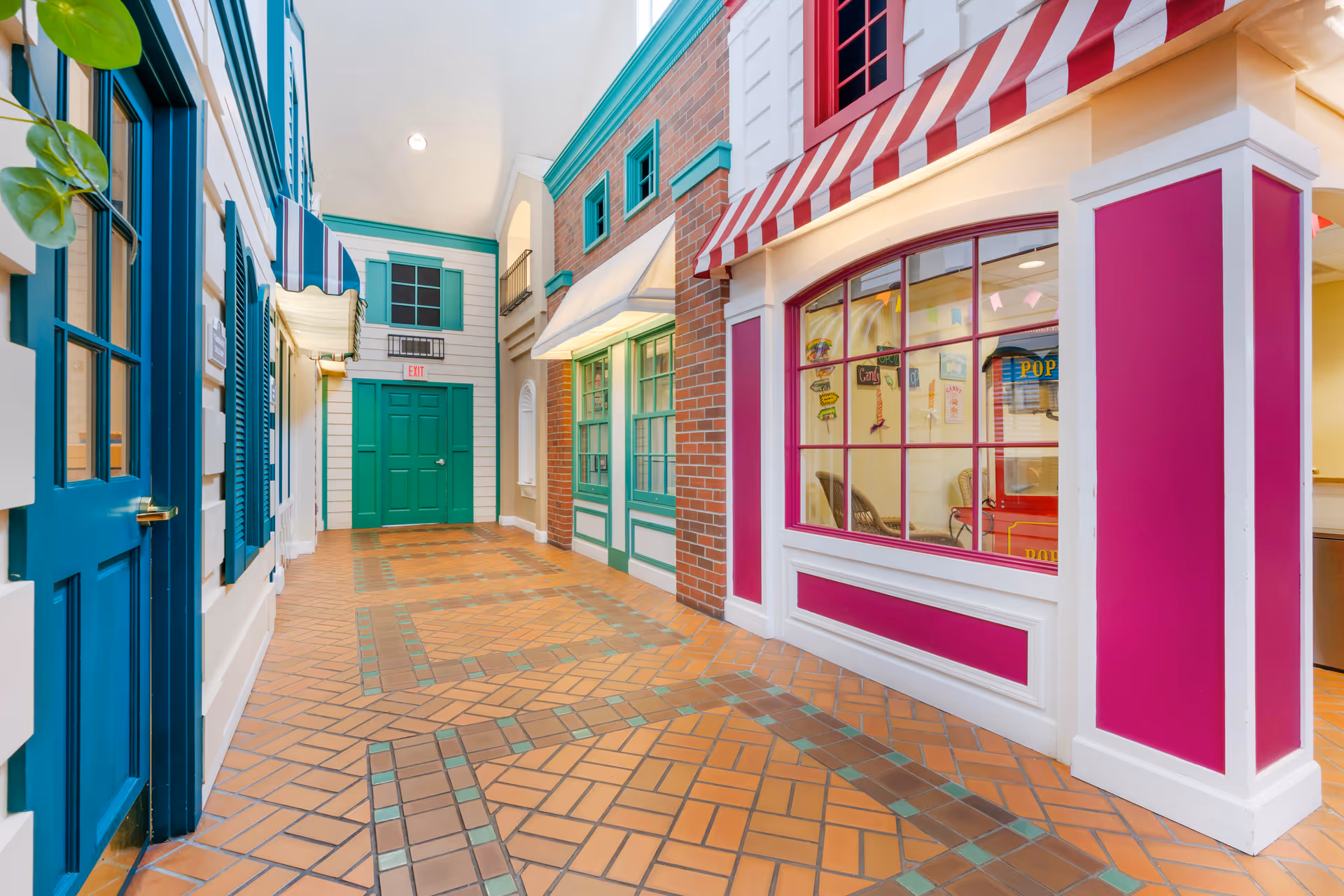 Indoor hallway designed to look like an outdoor street with colorful storefront-style walls, green and blue doors, windows with awnings, and a tiled floor with a geometric pattern.