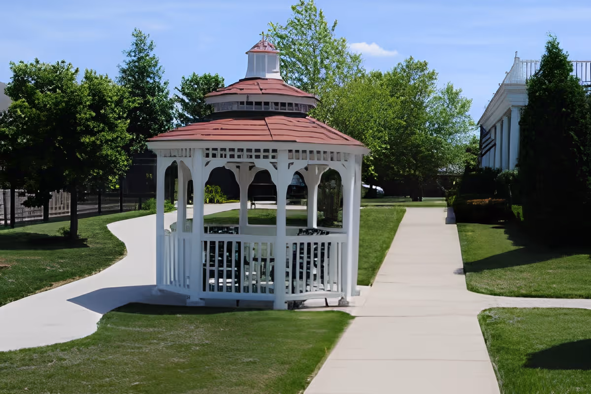 A white gazebo with a red roof situated on a green lawn with paved walkways leading around it. Trees and bushes surround the area, and a building with white columns is visible on the right side under a clear blue sky.