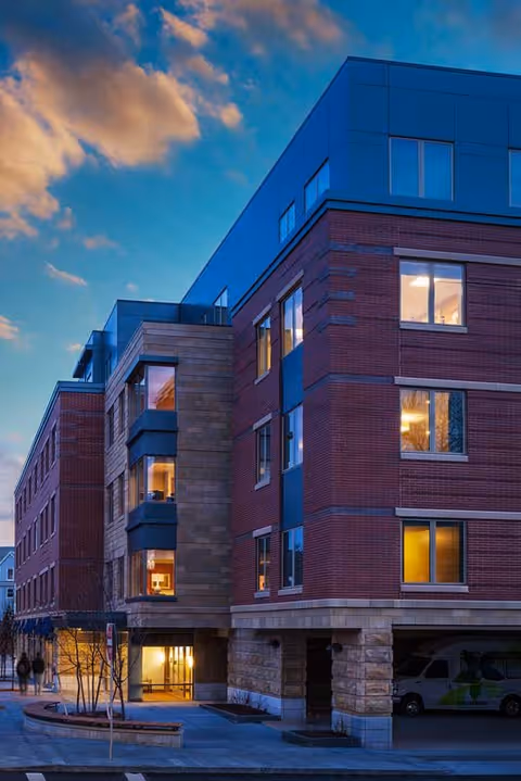 Exterior view of a modern multi-story residential building at dusk with warm lights glowing from the windows and a partly cloudy sky in the background.