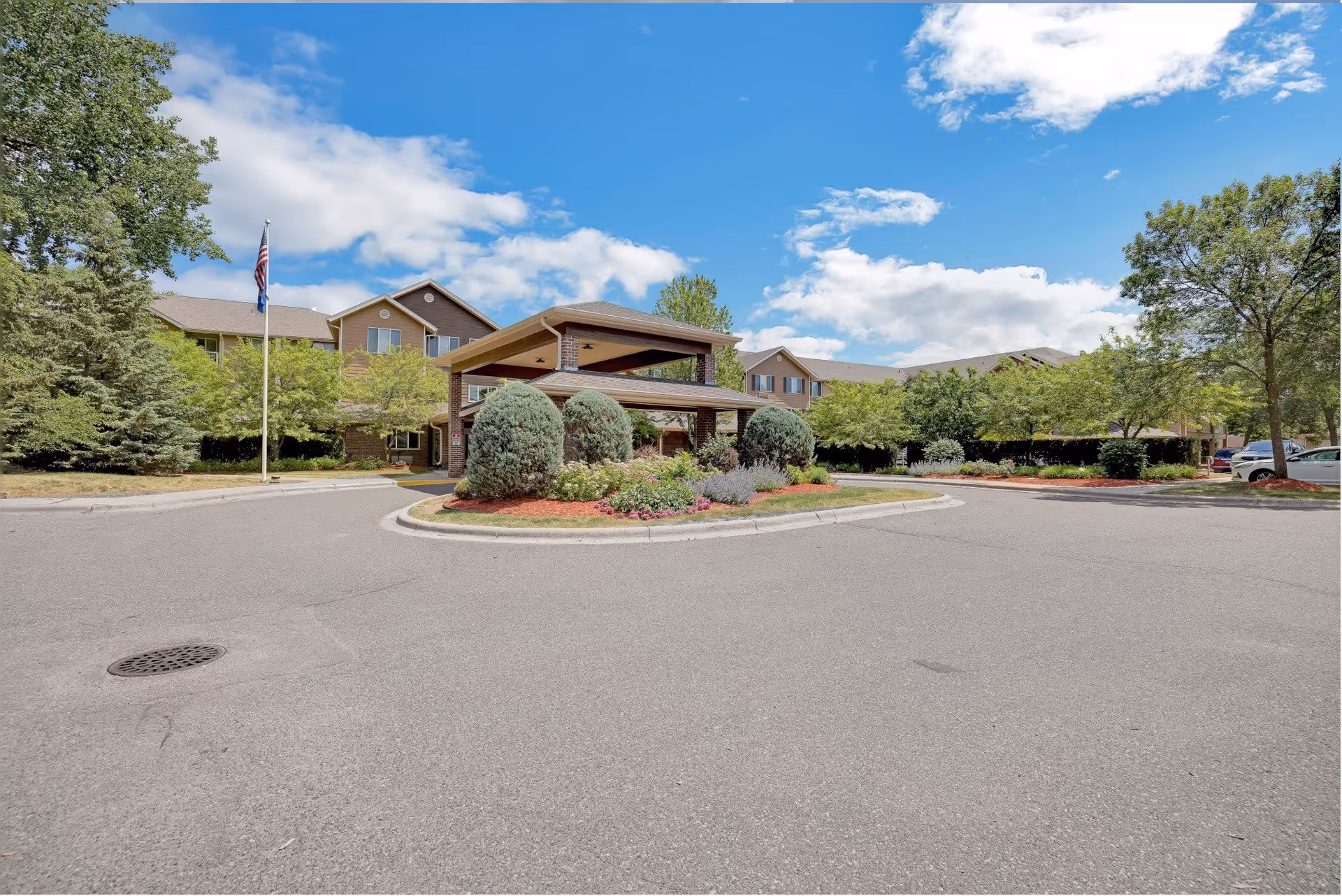 Exterior view of The Lodge at White Bear, a senior living community, showing a driveway with a landscaped roundabout featuring bushes and flowers. The building is a two-story structure with a covered entrance, surrounded by trees and parked cars under a partly cloudy blue sky.