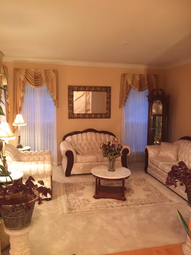 A cozy living room with beige walls and carpet, featuring a matching set of cream-colored upholstered sofas and armchairs with dark wooden accents. A round marble-top coffee table with a vase of flowers sits on a floral area rug. Two tall windows with sheer white curtains and decorative valances flank a wall with a large ornate mirror. A grandfather clock stands in the corner, and there are potted plants adding greenery to the space.