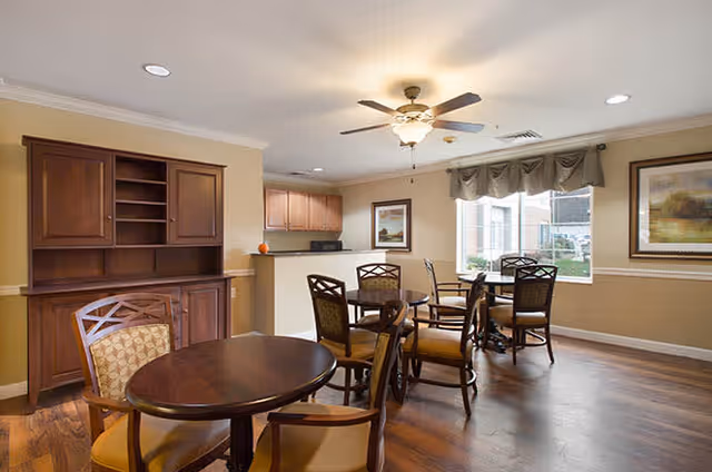 A well-lit common area with multiple round wooden tables and cushioned chairs arranged on a hardwood floor. There is a ceiling fan with a light fixture, a large window with a valance, framed artwork on the walls, and a wooden cabinet with shelves. The area appears clean and inviting, suitable for social gatherings or dining.