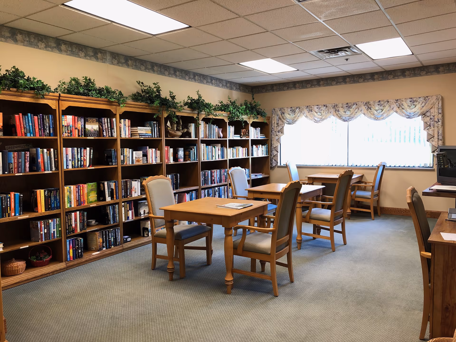 A quiet library or reading room with wooden bookshelves filled with books along one wall, several wooden tables with cushioned chairs, a large window with floral valance curtains letting in natural light, and a computer station on the right side.