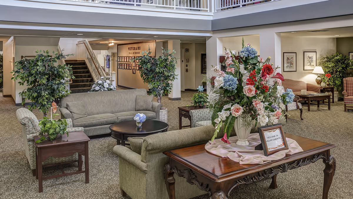 Spacious senior living lobby with upholstered sofas and chairs, tables, plants, and a large floral arrangement on a wooden table.