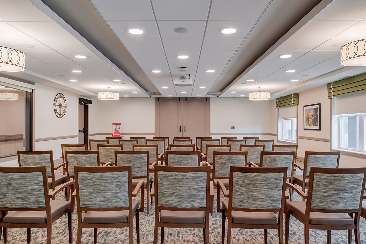A conference or meeting room with rows of wooden chairs facing a closed double door. The room has a carpeted floor, ceiling lights, a wall clock, a popcorn machine on a table, and windows with green valances and framed artwork on the walls.