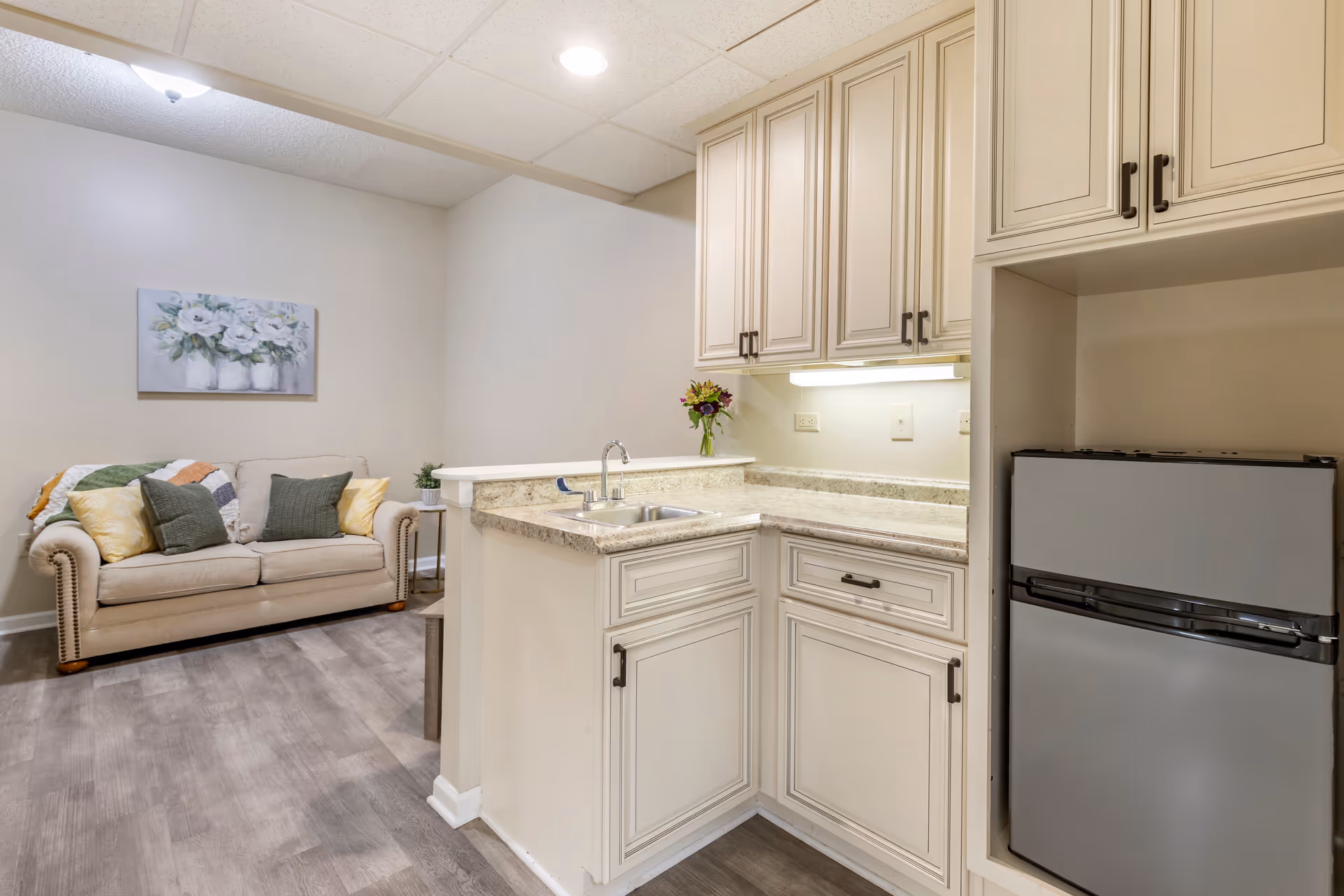 Small kitchenette with cream cabinets and a sink opening onto a living area with a beige sofa and decorative pillows.