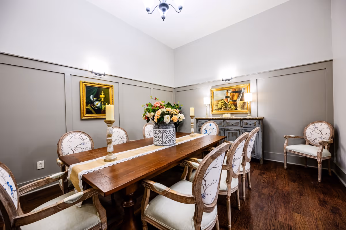 A well-lit dining room with a long wooden table surrounded by eight upholstered chairs with floral patterns. The table is decorated with a floral centerpiece and two tall candles. The walls are painted gray with white trim, and two framed paintings hang on the walls. A sideboard with two lamps is against the far wall, and the floor is dark hardwood.