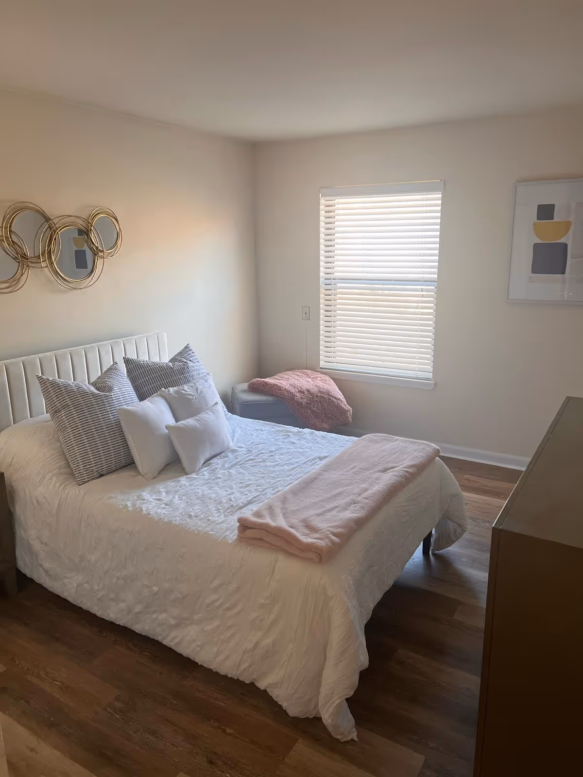 A neatly made bed with white bedding and multiple pillows in a bedroom with wooden flooring. There is a pink blanket folded at the foot of the bed. A window with white blinds is on the wall, and a small cushioned bench with a pink throw blanket is beneath it. Decorative circular mirrors are hung on the wall above the bed, and a framed abstract art piece is on the adjacent wall.
