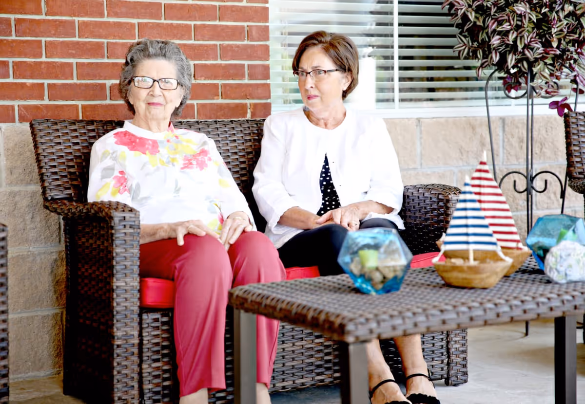 Two elderly women sitting on a wicker loveseat with red cushions on a patio. One woman is wearing glasses, a white floral top, and red pants, while the other woman is wearing glasses, a white jacket, and black pants. In front of them is a wicker table with decorative items including a small model sailboat and glass terrariums. The background shows a brick and stone wall with a window and blinds.