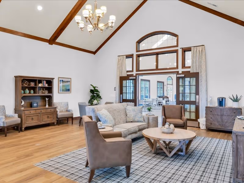 A spacious living room with high vaulted ceilings featuring wooden beams and a chandelier. The room includes a light gray sofa with blue pillows, two brown armchairs, a round wooden coffee table on a plaid area rug, and wooden furniture including a cabinet and sideboard. Large windows and glass doors with curtains allow natural light to enter, leading to another room with tables and chairs.