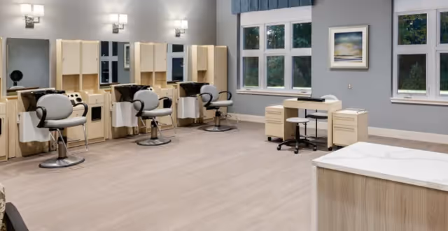 A spacious salon area with three styling stations, each equipped with a mirror, chair, and storage cabinets. The room has light wood flooring, gray walls, and large windows letting in natural light. There is also a small desk with a chair near the windows and a framed picture on the wall.