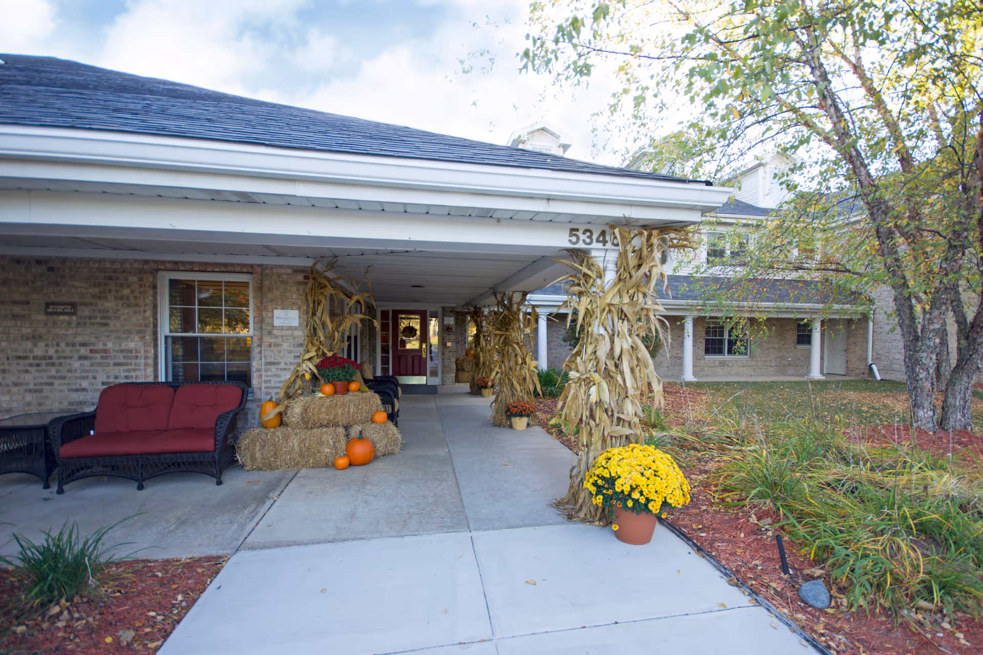 Entrance of Brookdale Meridian - Assisted Living decorated for fall with hay bales, pumpkins, corn stalks, and potted yellow flowers. There is a covered walkway with a red cushioned bench on the left and a brick building in the background.