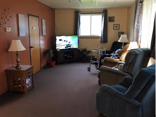 A cozy living room with brown carpet and walls painted in beige and burnt orange. The room features a wooden side table with a lamp, a flat-screen TV on a wooden stand, a walker, and several cushioned chairs and recliners. There are two windows with curtains allowing natural light to enter the room.
