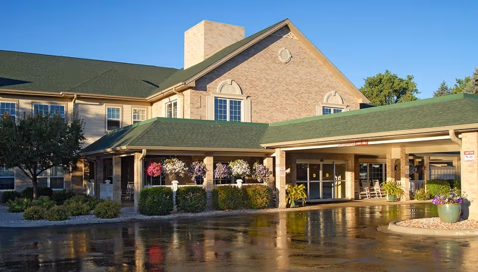 Exterior view of a senior living facility building with beige brick walls and a green roof. The entrance has a covered driveway with hanging flower baskets and potted plants. There are several windows and a tree with shrubs in front of the building. The pavement is wet, reflecting the building and plants.