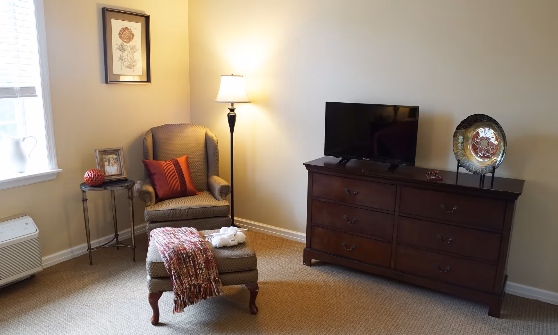 A cozy living room corner with a cushioned armchair and matching ottoman draped with a red and white throw blanket. A red pillow is on the armchair. Next to the chair is a small round side table with a framed photo and a decorative red object. A floor lamp provides warm lighting. Across from the chair is a wooden dresser with a flat-screen TV and a decorative plate on a stand. The room has beige walls and carpeted floor, with a window letting in natural light.