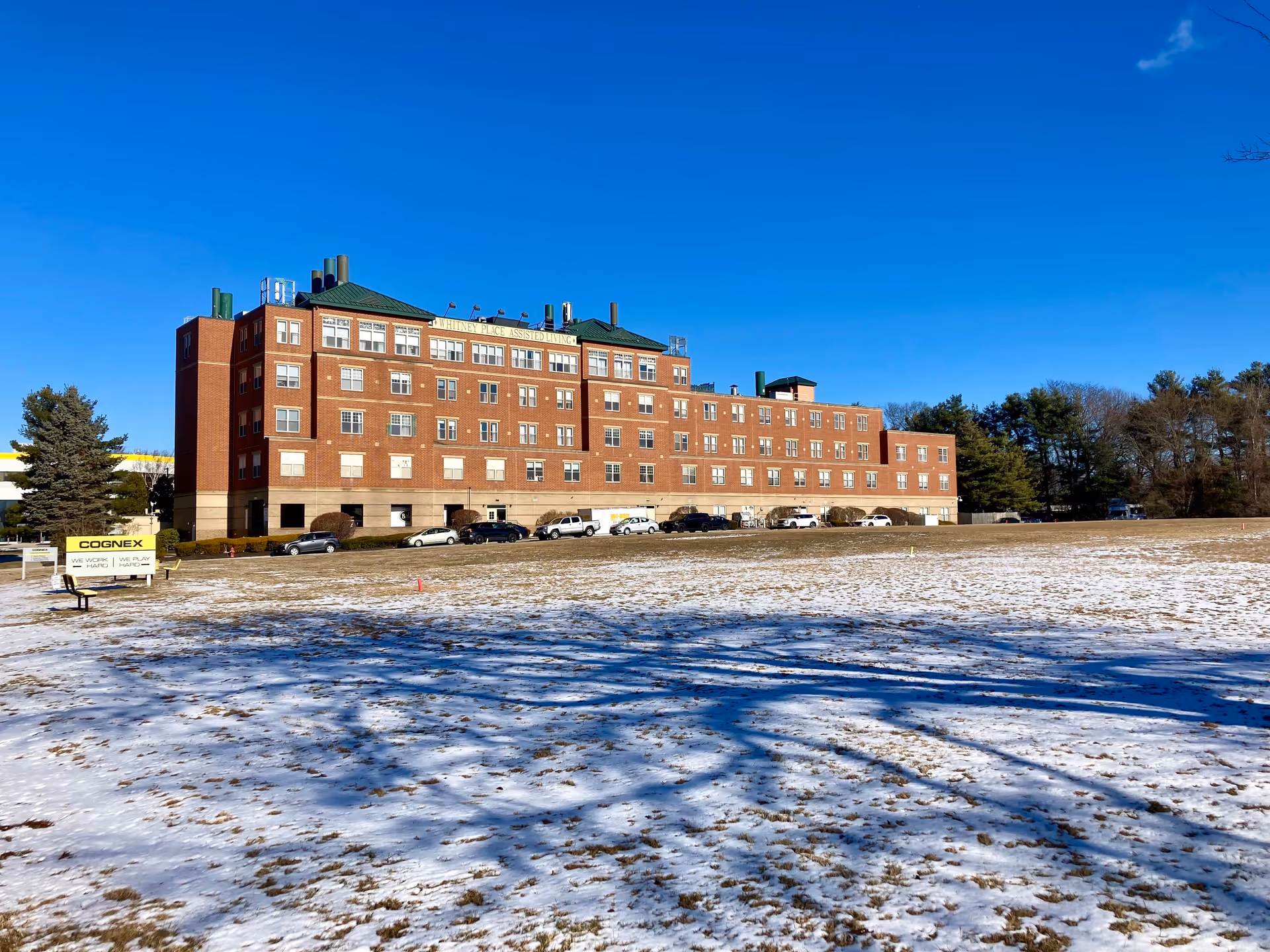A large multi-story brick building under a clear blue sky with a snow-covered grassy field in the foreground. Several cars are parked along the side of the building, which has multiple windows and green roof accents. Trees are visible in the background.