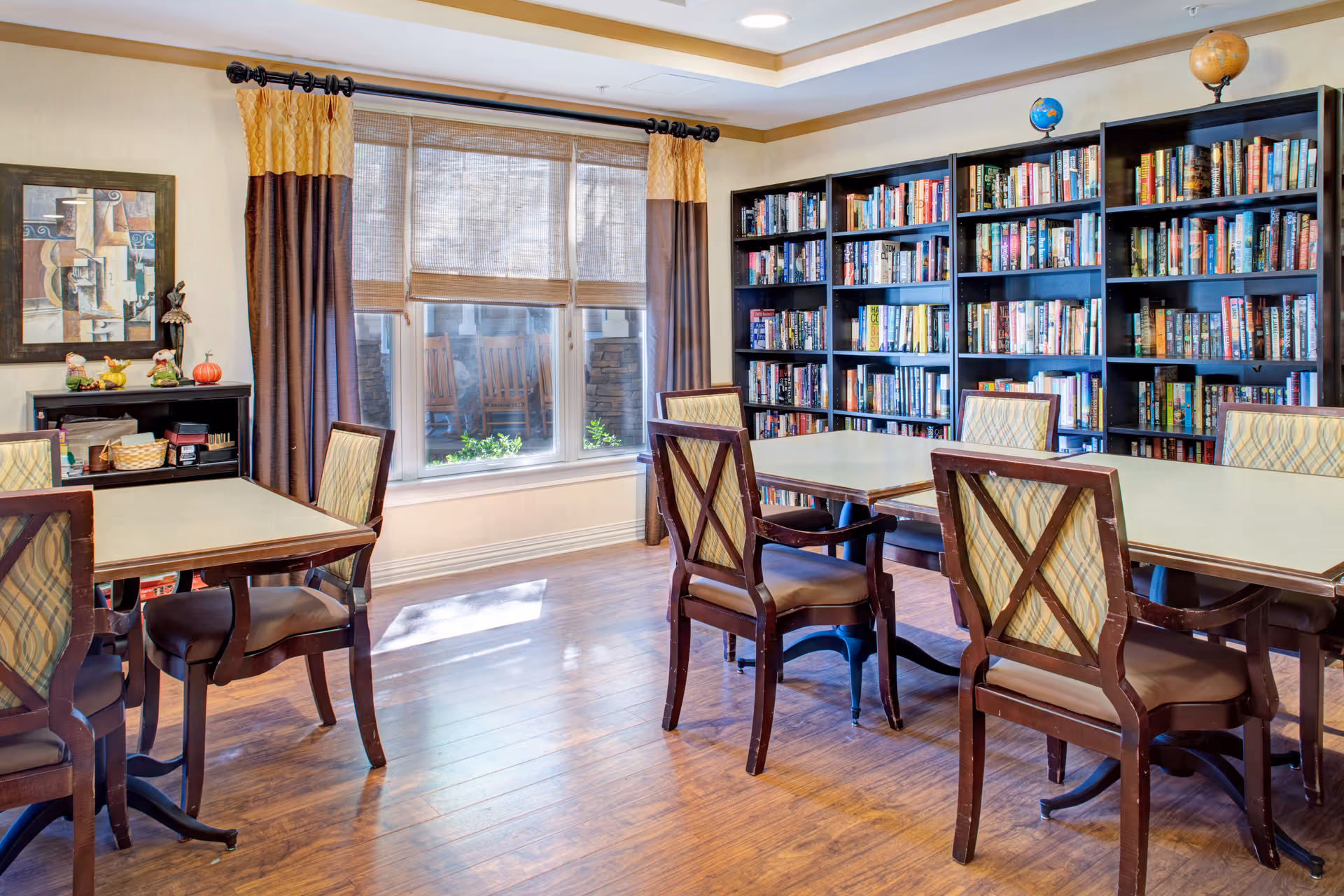 A well-lit room with wooden flooring featuring several tables and chairs arranged for seating. A large window with beige and brown curtains allows natural light to enter. Along one wall, there are multiple bookshelves filled with books. A small cabinet with decorative items and a framed abstract painting is also visible.