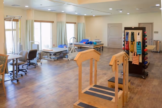 A spacious rehabilitation therapy room with wooden parallel bars in the foreground, therapy tables with blue and red cushions, chairs, and various exercise equipment. Large windows with curtains allow natural light to fill the room.