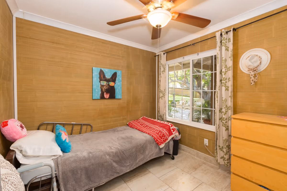 Small single bedroom with a twin bed, ceiling fan, window with curtains, wooden dresser, and a dog painting on the wall.