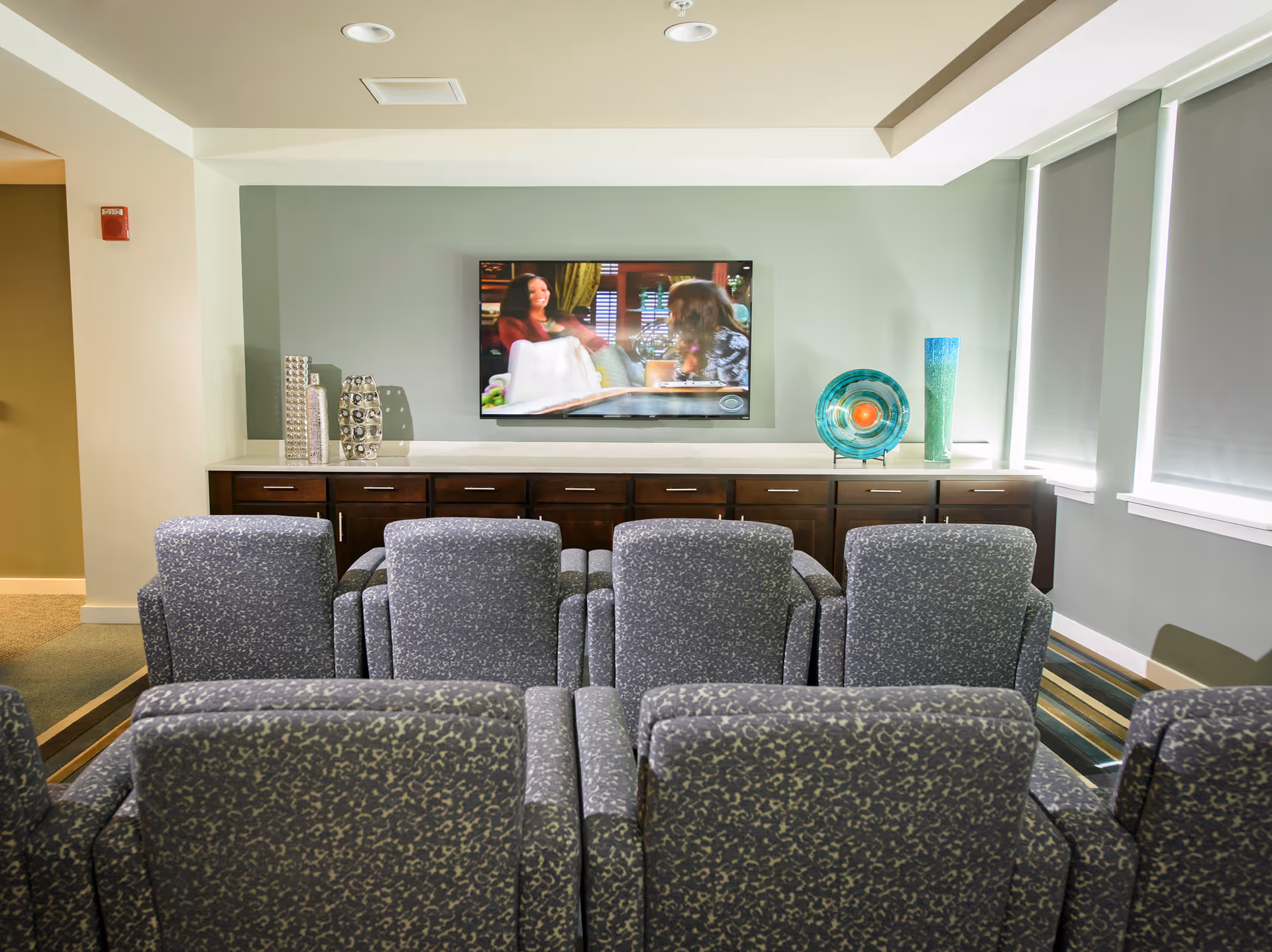 A small theater-style room with two rows of patterned gray armchairs facing a wall-mounted flat-screen TV. Below the TV is a long wooden cabinet with decorative vases and a colorful glass plate. The room has light green walls and windows with gray blinds on the right side.