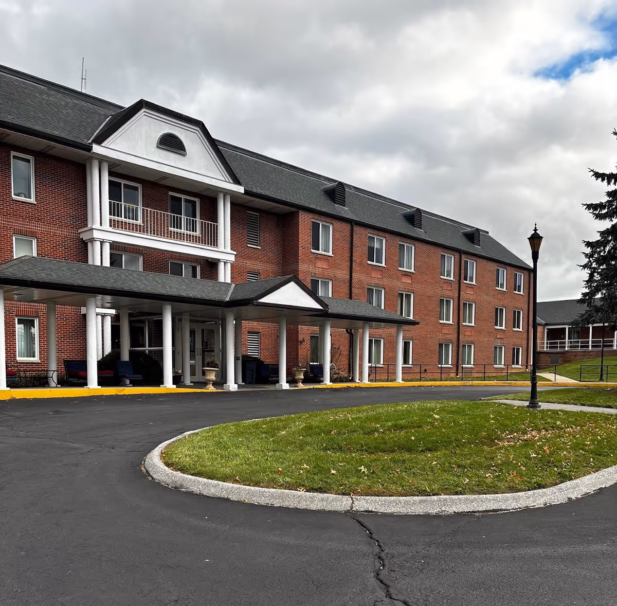 Exterior view of a three-story brick senior living facility with a covered entrance supported by white columns, a circular driveway with a grassy island in the center, and a lamppost on the right side under a cloudy sky.