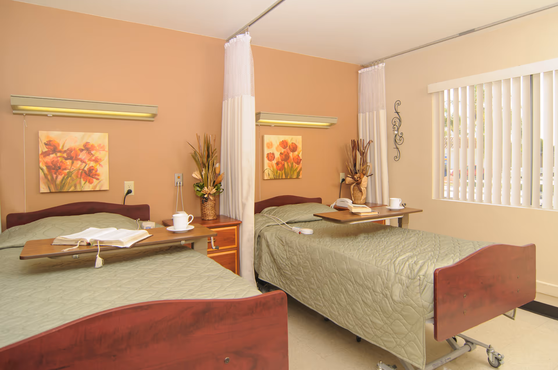 A hospital room with two beds separated by a privacy curtain. Each bed has a green quilted blanket, a wooden overbed table with a cup and book, and a floral painting on the wall above the headboard. There is a window with vertical blinds on the right side, and decorative plants on the nightstand between the beds.