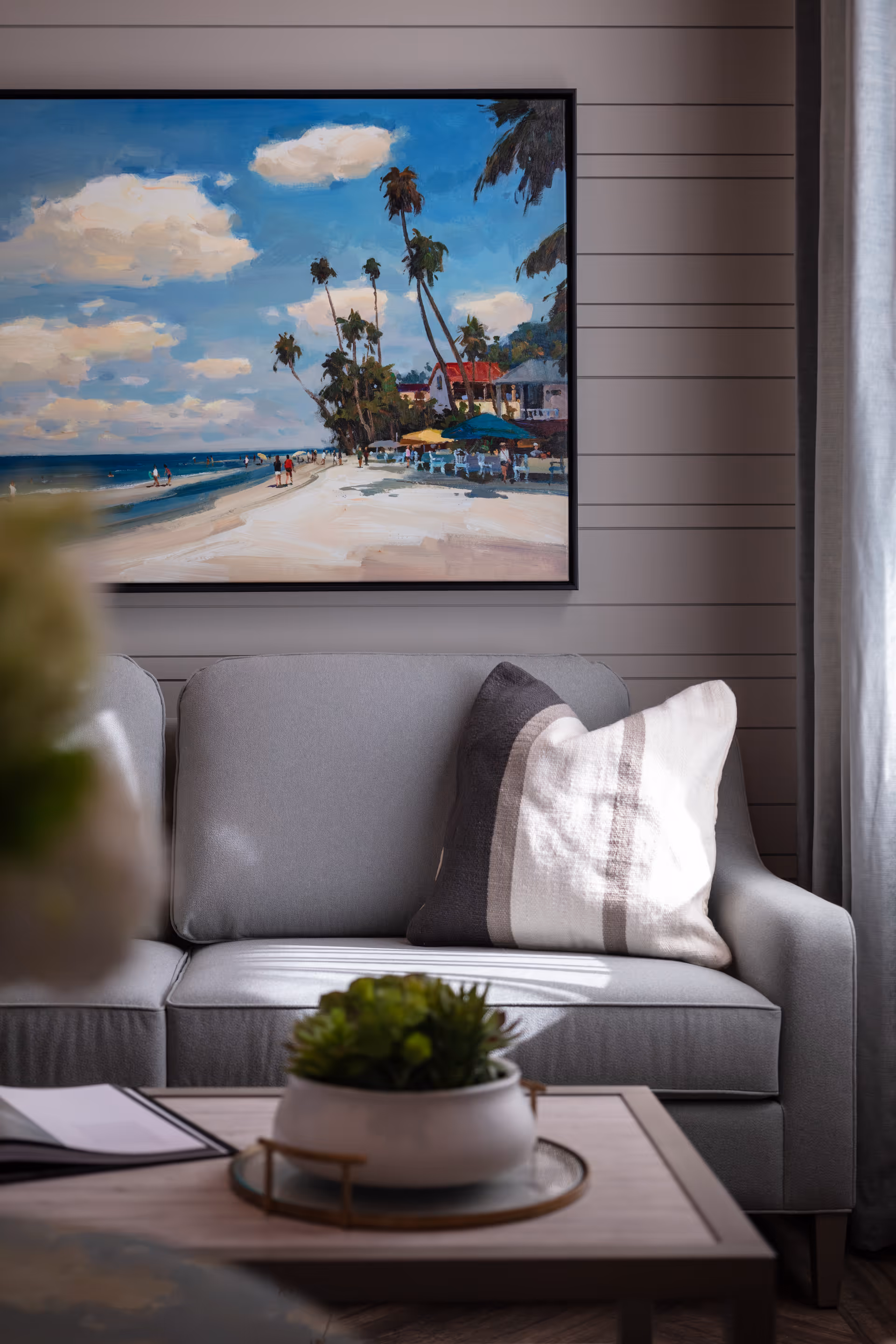 A cozy living room area with a light gray sofa adorned with a striped pillow. Above the sofa hangs a framed painting depicting a beach scene with palm trees, people, and colorful umbrellas under a partly cloudy sky. In front of the sofa is a wooden coffee table with a small potted plant and an open book or magazine. Light filters in from a window with gray curtains on the right side.