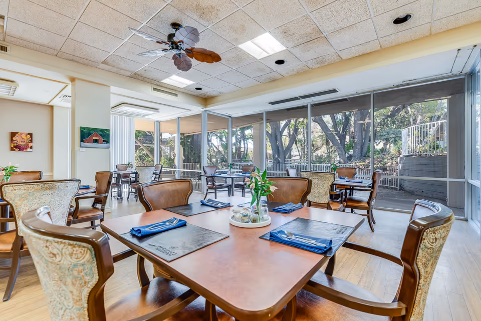 A bright dining room with several wooden tables and chairs, each table set with blue napkins and menus. Large windows on one side provide a view of trees and outdoor greenery. The ceiling has a fan with leaf-shaped blades, and there are paintings on the walls.