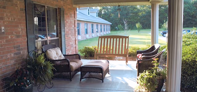 Covered outdoor patio area with wicker furniture including a cushioned chair, ottoman, two chairs, and a wooden bench swing. The patio overlooks a grassy yard with trees and a brick building in the background.
