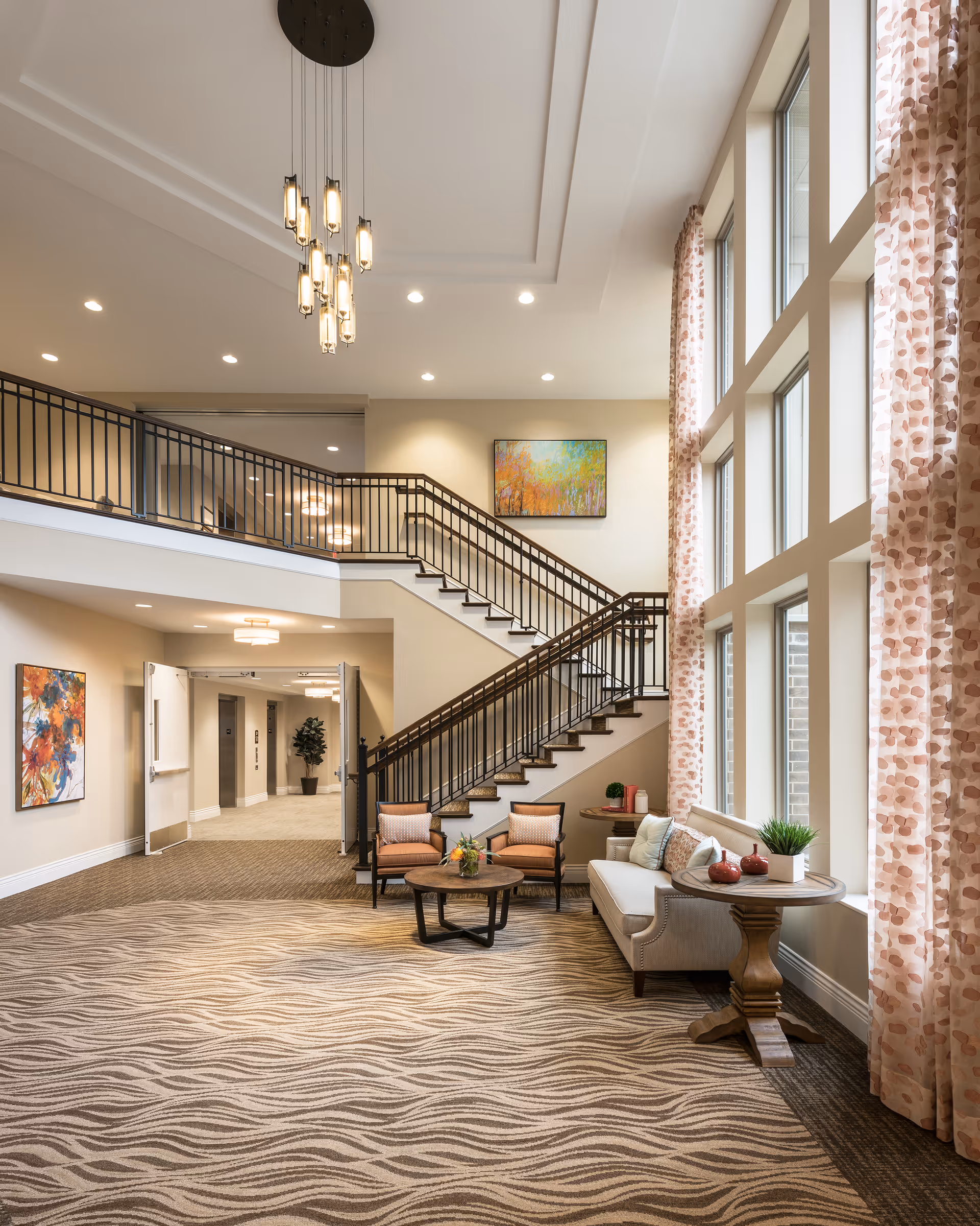 A spacious and well-lit interior common area with large windows covered by patterned curtains, a beige sofa with cushions, two armchairs, a round wooden coffee table with a flower arrangement, and a side table with decorative items. A staircase with black railings leads to an upper floor, and colorful paintings hang on the walls. The carpet has a wavy pattern, and modern ceiling lights illuminate the space.