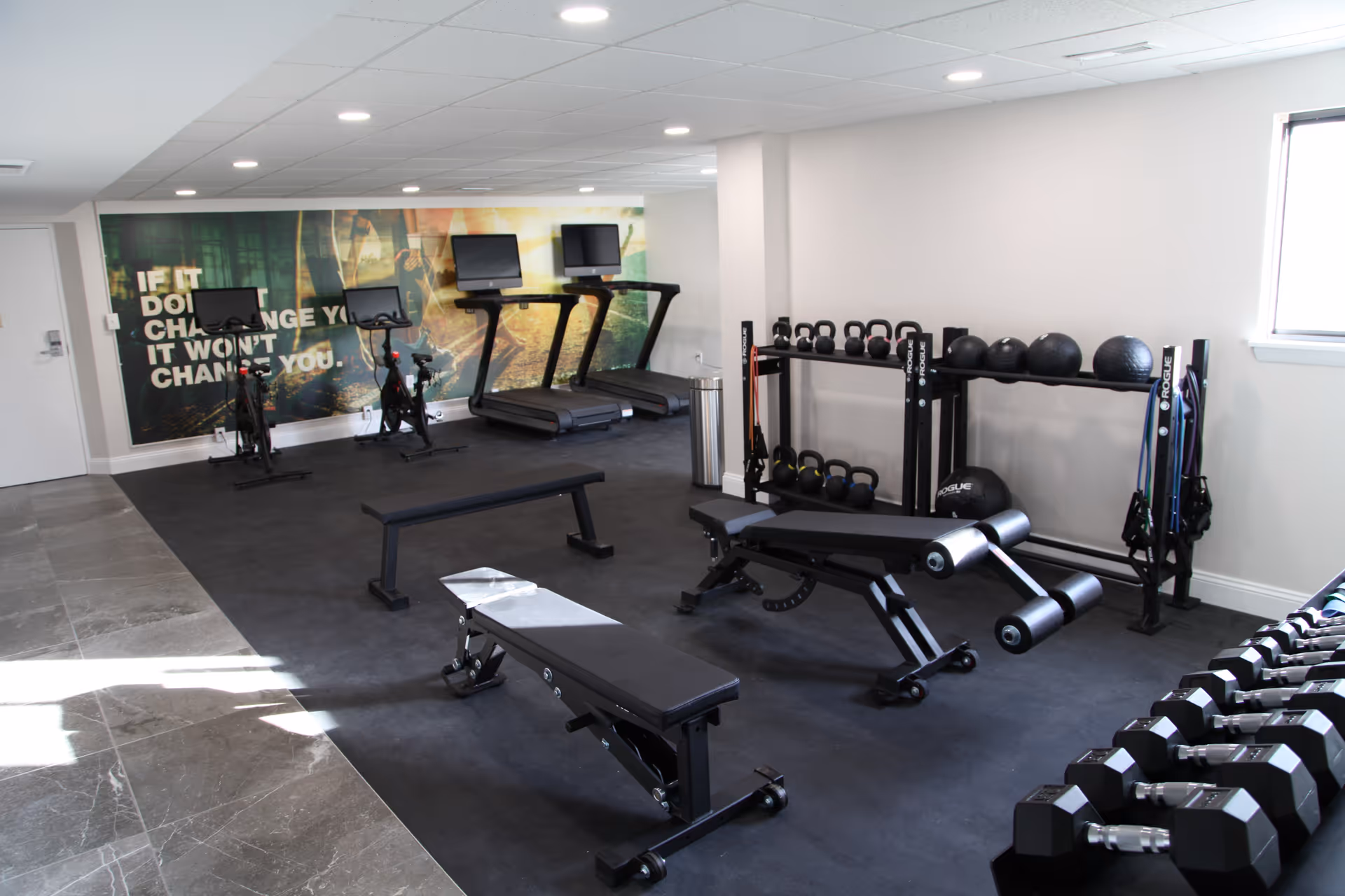A modern fitness room with exercise equipment including two stationary bikes, two treadmills, weight benches, dumbbells, kettlebells, medicine balls, and resistance bands. The room has a large motivational wall mural that reads 'IF IT DOESN'T CHANGE YOU IT WON'T CHANGE YOU.'